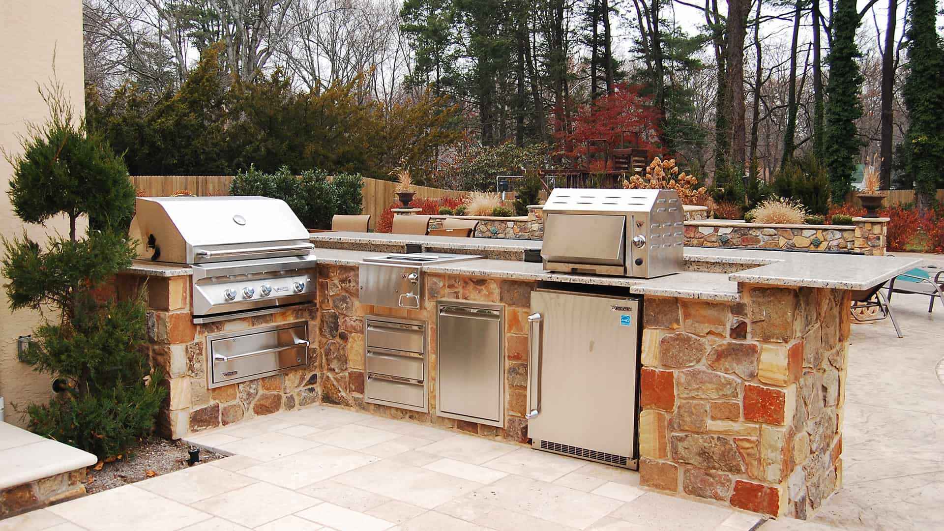 Discover the perfect outdoor living space in Delaware County, PA, with a stone-counter kitchen featuring two stainless steel grills and storage cabinets. Nestled among trees with a patio area, this setup offers a cozy and functional environment for alfresco cooking.