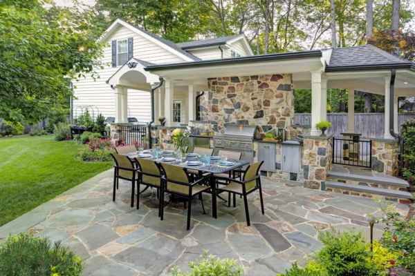 Outdoor patio with a dining table set for eight on a stone floor, an inviting space that showcases the essence of outdoor living in Delaware County, PA. A built-in grill and countertop are under a covered area with stone walls, while lush greenery and flowers adorn the landscaped yard beside a partially visible white house.