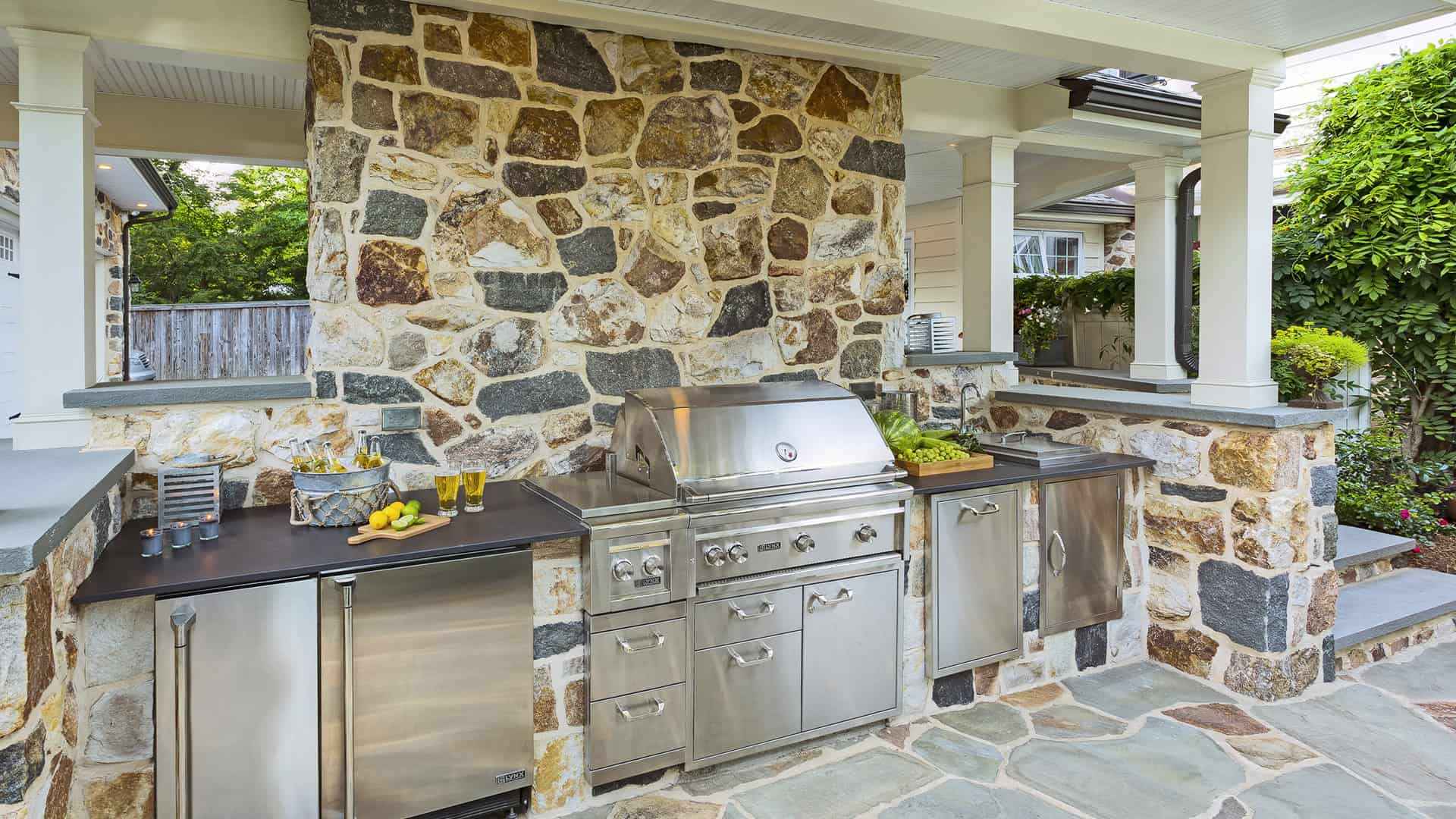 Outdoor kitchen with stone walls featuring a stainless steel grill, refrigerator, and storage. A countertop holds beverages, lemons, and a salad bowl. Surrounded by plants and patio space under a white ceiling, showcasing the finest in outdoor living Delaware County PA.