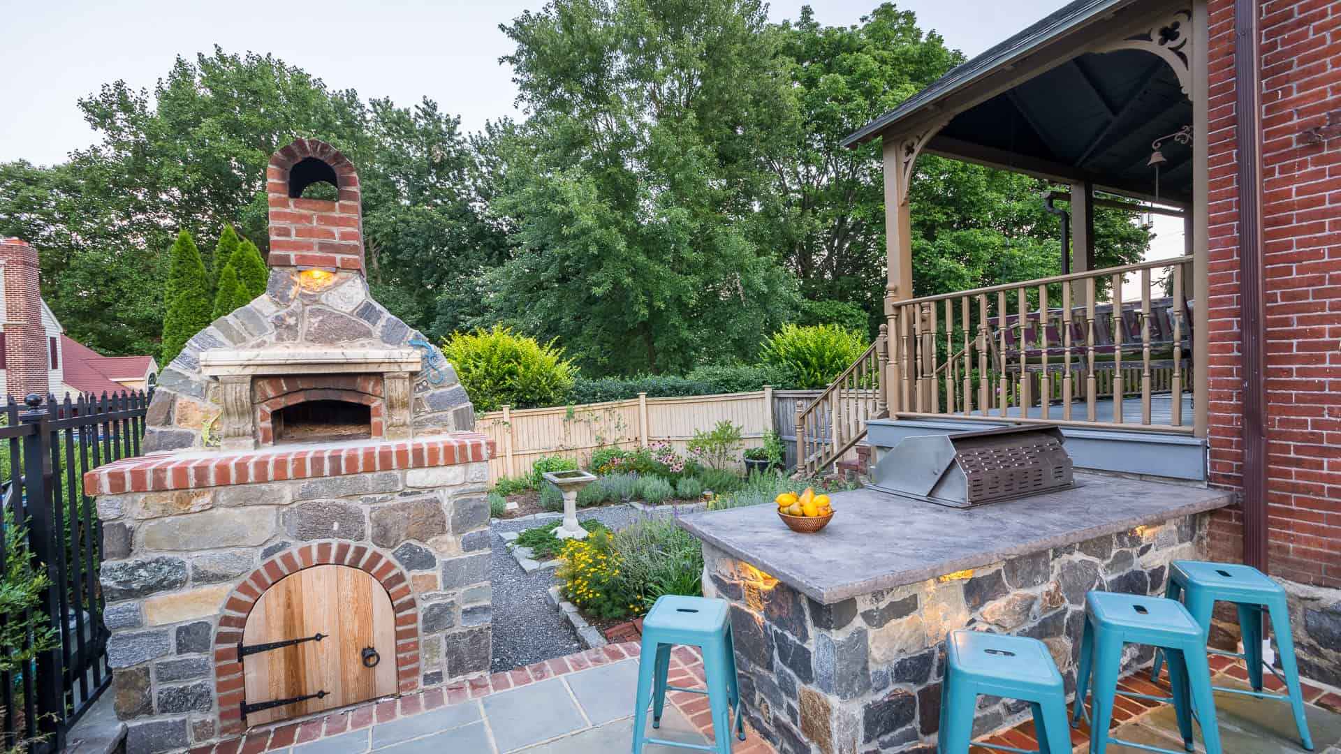 Outdoor patio with a stone brick pizza oven, adjacent countertop with four blue stools, and a bowl of oranges. Behind is a wooden fence, lush greenery, and part of a brick house with a covered porch—perfect for those enjoying outdoor living in Delaware County, PA.