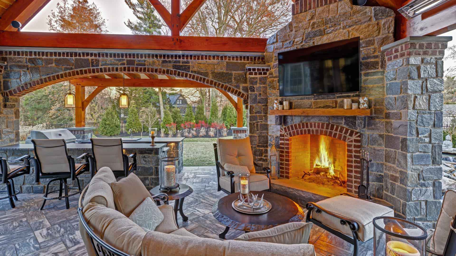 Outdoor living in Delaware County, PA is exemplified by this patio with a stone fireplace, cozy seating, and a TV under a wooden pergola. A bar with stools sits to the left. Surrounding trees and manicured gardens enhance the inviting atmosphere.
