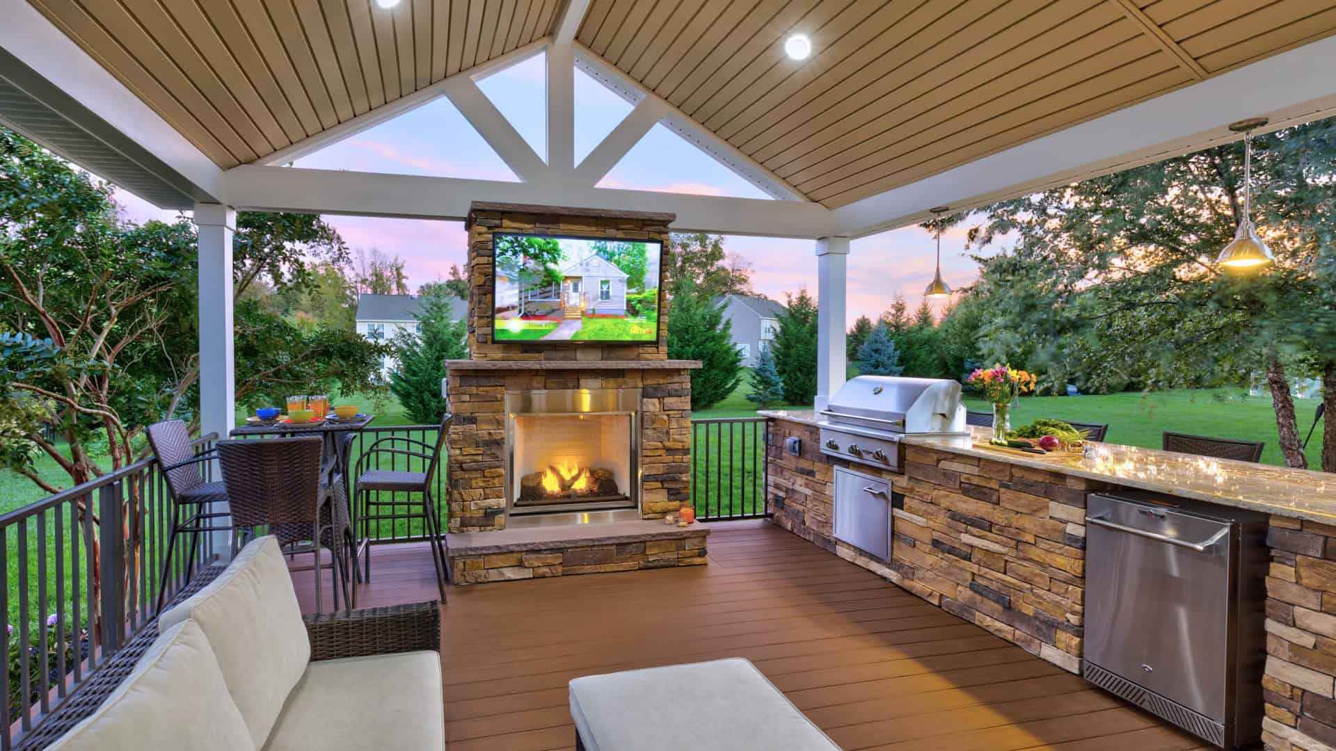 Outdoor patio with a wooden deck and covered roof, perfect for outdoor living in Delaware County, PA. Features a stone fireplace with a TV above, grill, refrigerator, and a bar area. Comfortable seating and dining table amidst lush greenery and a sunset sky complete the scene.