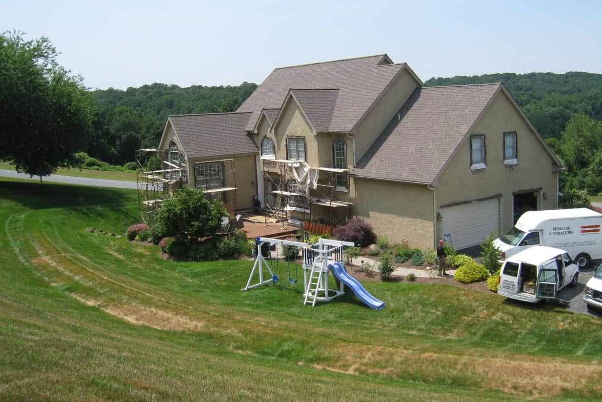 A beige two-story house is under construction in Delaware County, PA, with scaffolding on the facade. A blue and white slide and swing set grace the backyard. Two white vans are parked in the driveway, amidst a large, grassy lawn and trees—a perfect setting for future outdoor living and landscaping services.