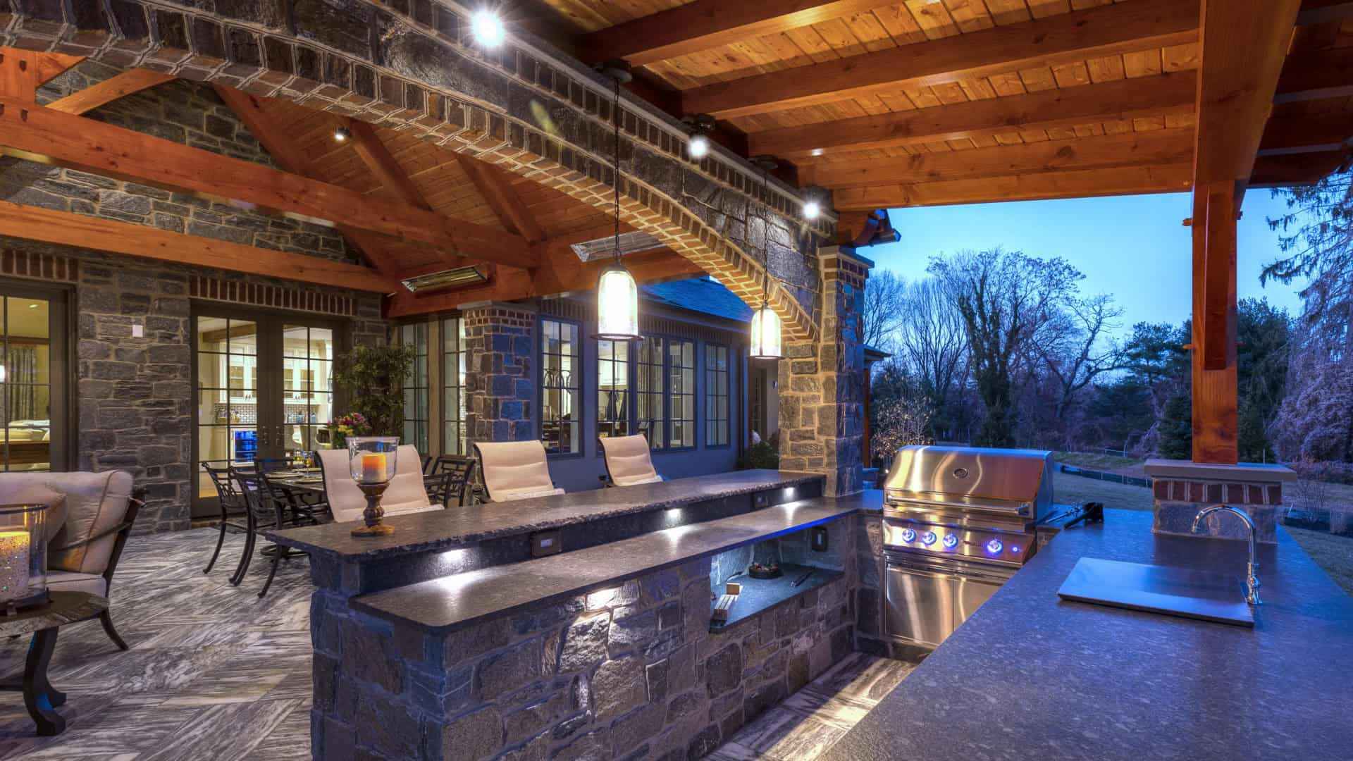 Outdoor living in Delaware County, PA comes to life with a kitchen featuring a stone and wood patio, built-in grill, countertop bar seating, and ambient lighting. A dining table and seating area sit under the twilight sky, surrounded by trees for the perfect outdoor escape.