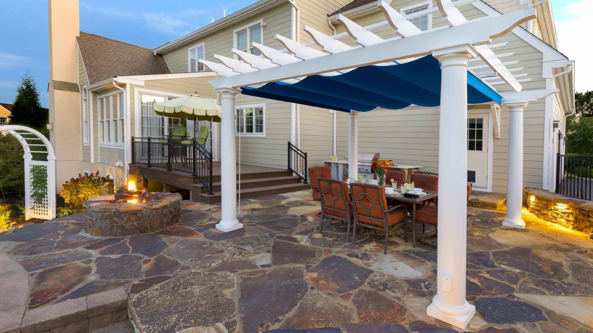 A backyard patio showcases a stone fire pit next to a dining set under a white pergola with a blue canopy. The house features beige siding and a staircase leading to a porch, complete with a green umbrella. Expert outdoor living in Delaware County, PA, ensures the evening lighting creates warmth and charm.