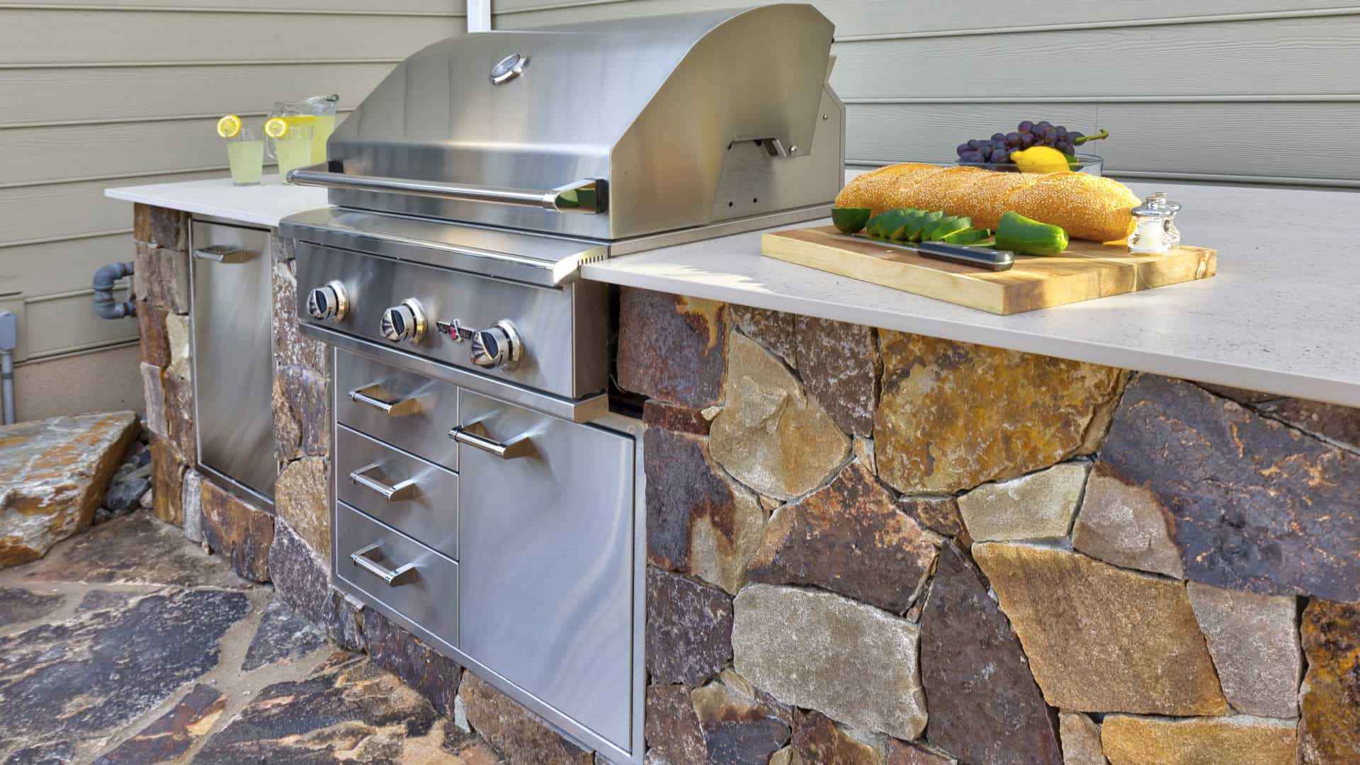 Outdoor kitchen with a stone base and stainless steel grill exemplifies outdoor living in Delaware County, PA. The countertop holds a cutting board with cucumbers, a baguette, cheese, and grapes. Nearby, a pitcher of lemonade is ready to enjoy on the beautifully crafted stone patio.