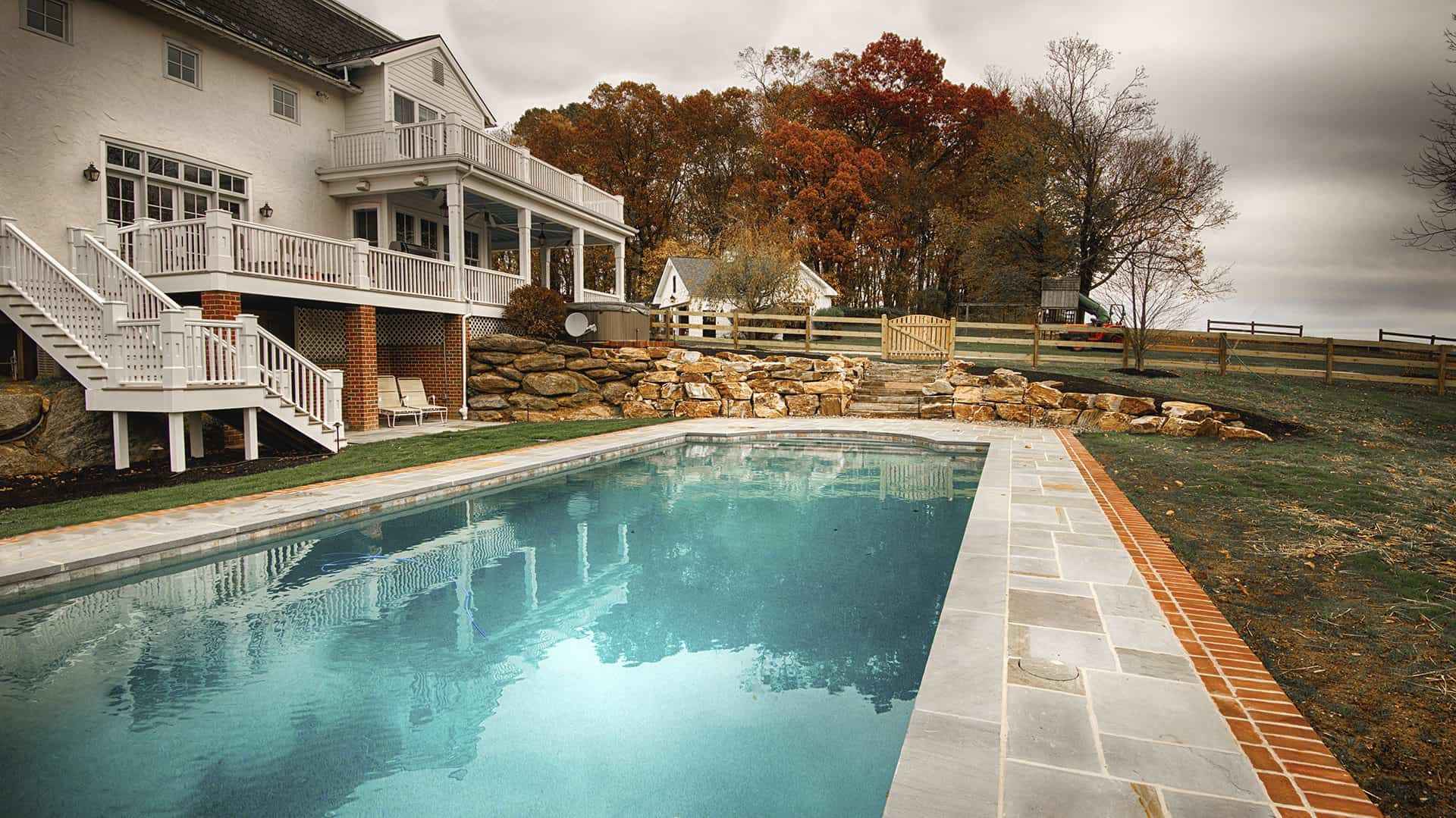 The backyard features a rectangular swimming pool framed by exquisite stone and brickwork, perfect for enhancing outdoor living in Delaware County, PA. The white house boasts a balcony with stairs leading down amid autumn trees and colorful leaves under a cloudy sky.