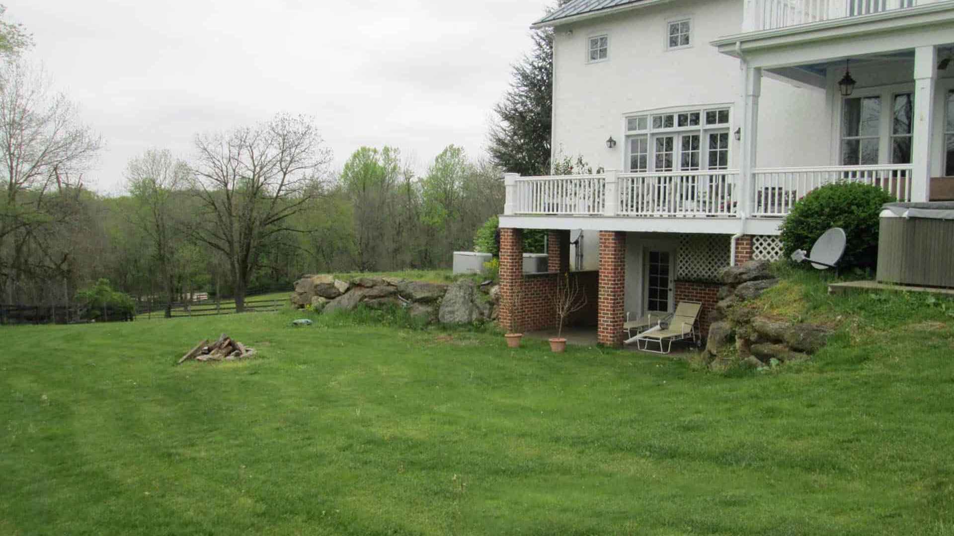 Large white house with a chimney and spacious lawn in Delaware County, PA. A small fire pit sits on the grassy area, blending perfectly with expertly designed hardscaping services. The building features a balcony, patio with lawn chairs and a satellite dish beneath it. Trees and a fence line the background under an overcast sky.