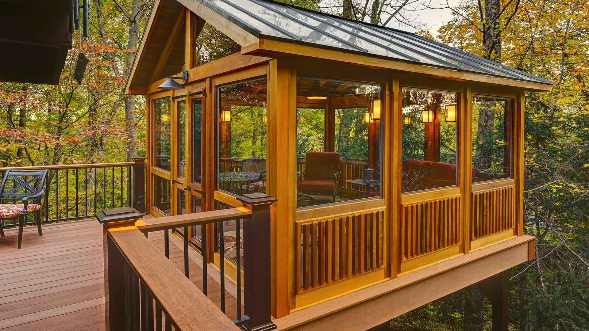 A cozy wooden sunroom with large windows extends from a house, surrounded by lush green trees. The interior features comfortable seating and warm lighting, making it a perfect outdoor living retreat in Delaware County, PA.