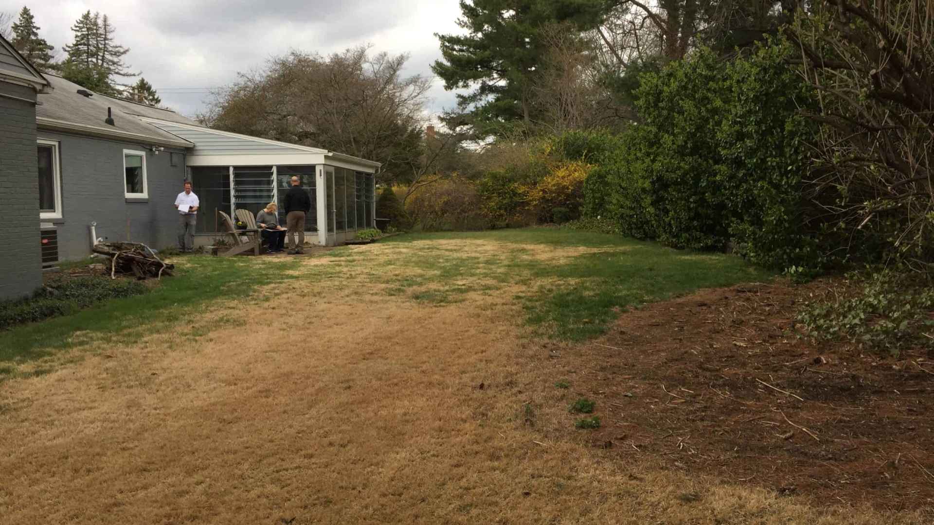 A backyard in Delaware County, PA, features a patchy lawn surrounded by trees and bushes. Two people stand near a white house with a glass-enclosed porch. A small stack of firewood rests against the house under the overcast sky, highlighting the potential for landscaping services.