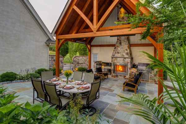 Outdoor living in Delaware County, PA, shines with a stone fireplace under a wooden pergola. A dining table for six is elegantly set, complemented by a nearby barbecue grill surrounded by lush greenery and trees. Rustic stone flooring showcases expert hardscaping services.