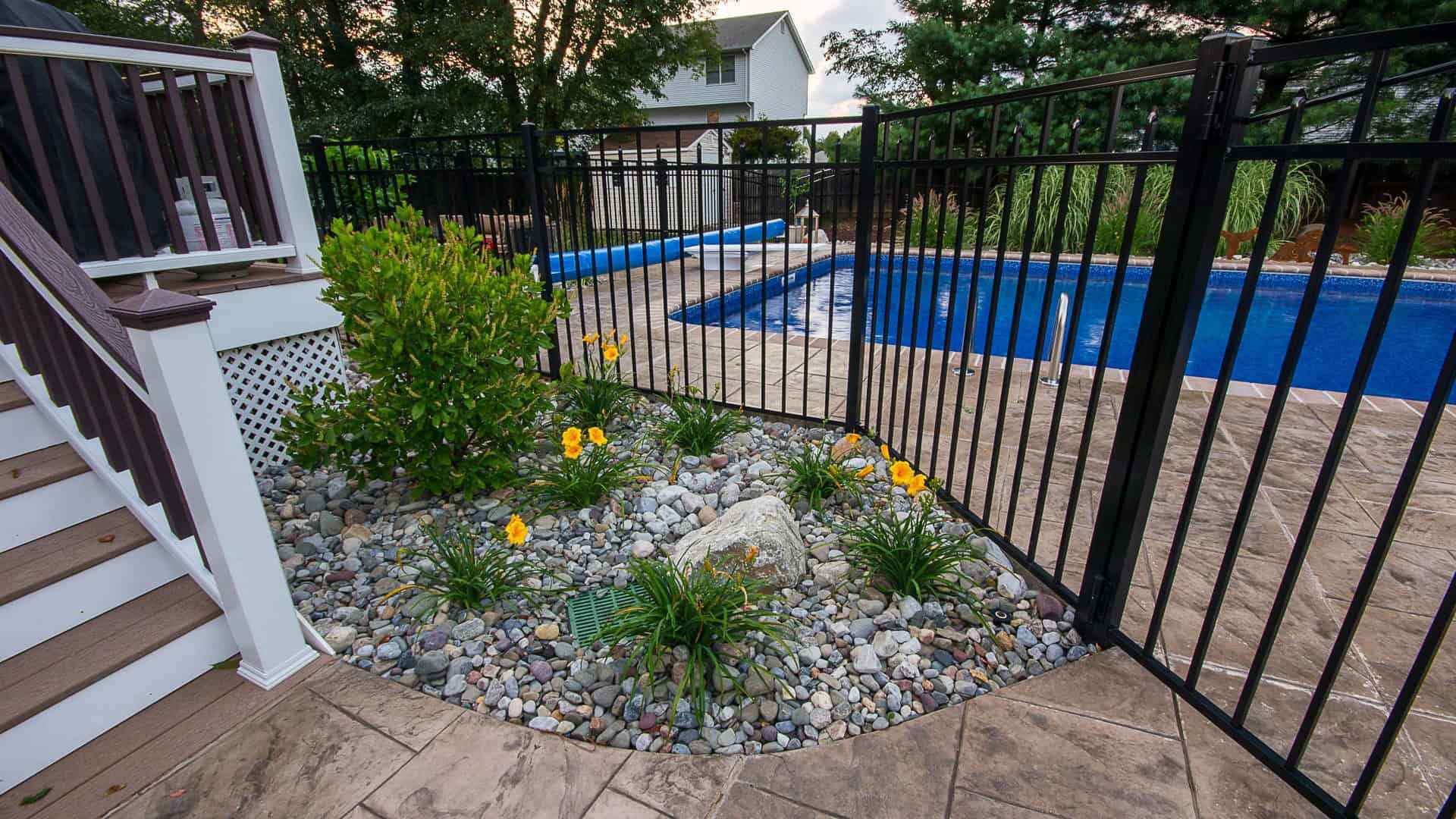 A fenced-in backyard pool area with a paved path showcases the epitome of outdoor living in Delaware County, PA. Yellow flowers and green shrubs thrive in a bed of small rocks. Stairs with a white railing lead up to a deck, framed by trees and a house in the background.