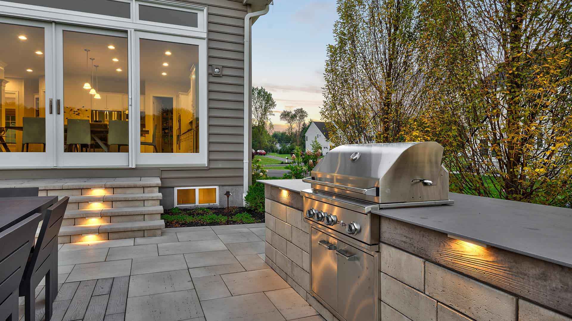Modern backyard patio in Delaware County, PA, featuring a stainless steel grill on a stone countertop. Warm lights accentuate the grill area. Nearby, a dining table with chairs awaits. Large glass doors lead to an illuminated interior space with a dining room, perfect for outdoor living enthusiasts.