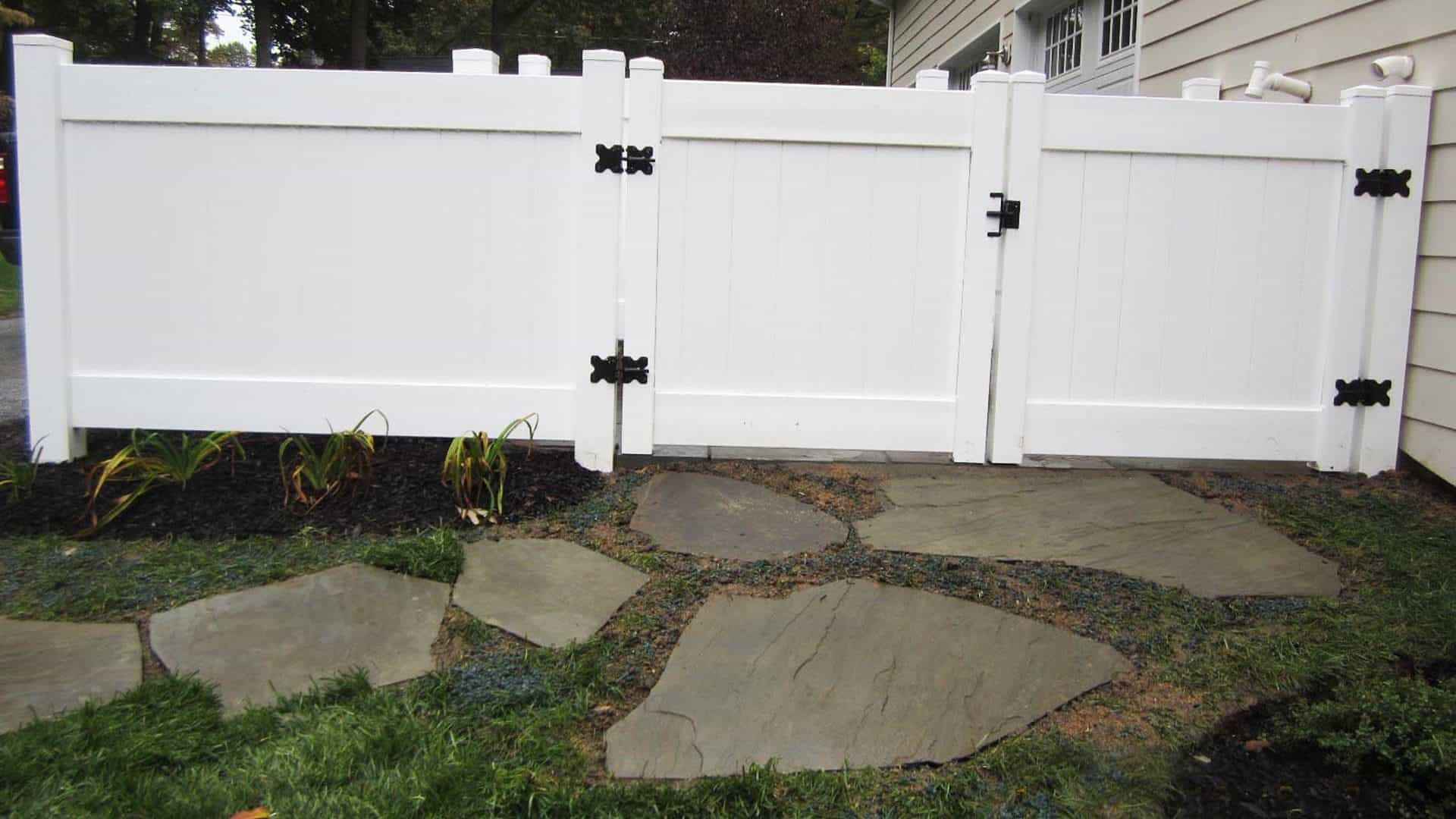 White vinyl fence with black hinges beside a beige house, featuring two gates on a blue-gray stone pathway lined with mulch and plants. Enhance your outdoor living in Delaware County, PA, with professional landscaping services. Trees add a natural backdrop to the serene scene.