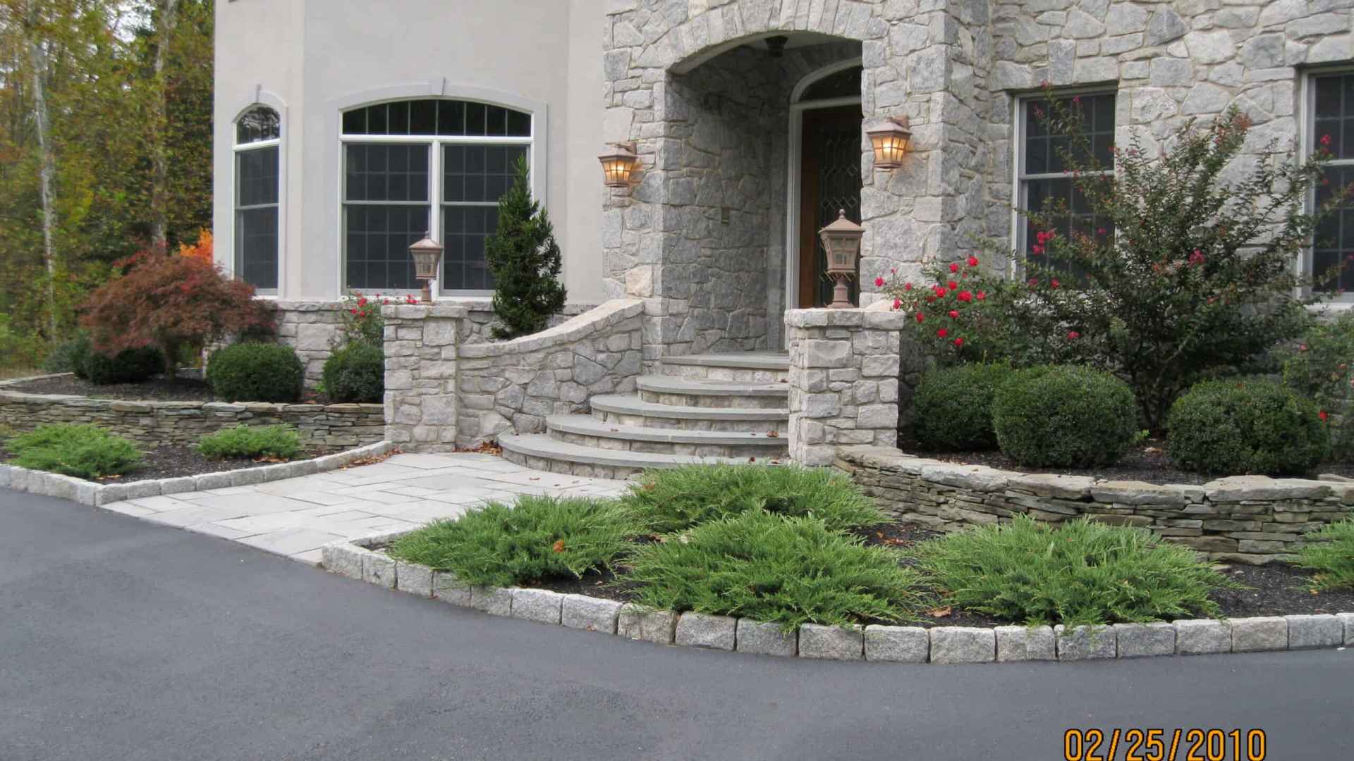 A stone house facade with an arched entryway and steps, flanked by two lit lanterns, showcases expert landscaping services in Delaware County, PA. The scene features shrubs, ornamental grasses, a stone retaining wall, two large windows, and a paved driveway in the foreground.
