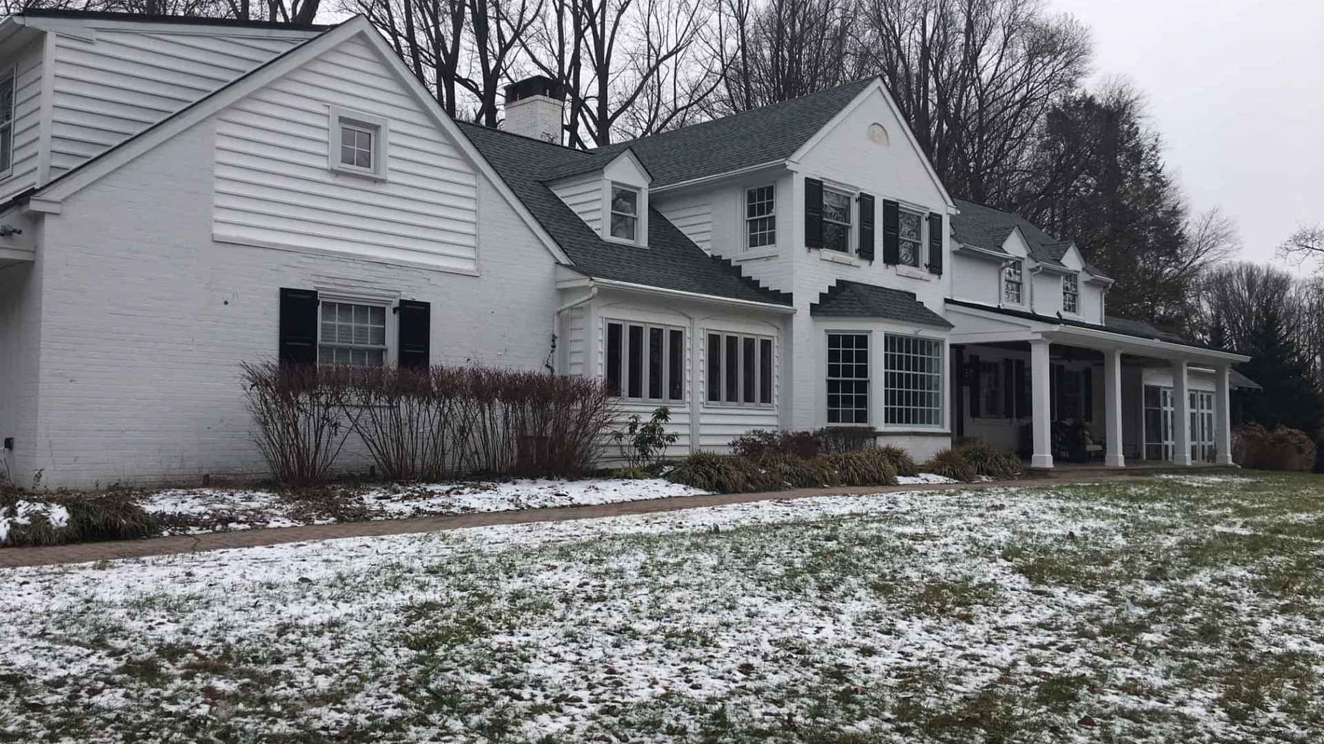 A large white house with black shutters and a gray roof stands amidst a snowy landscape, showcasing exceptional outdoor living in Delaware County, PA. Leafless trees are in the background, and a patchy snow-covered lawn is in the foreground. The sky is overcast.