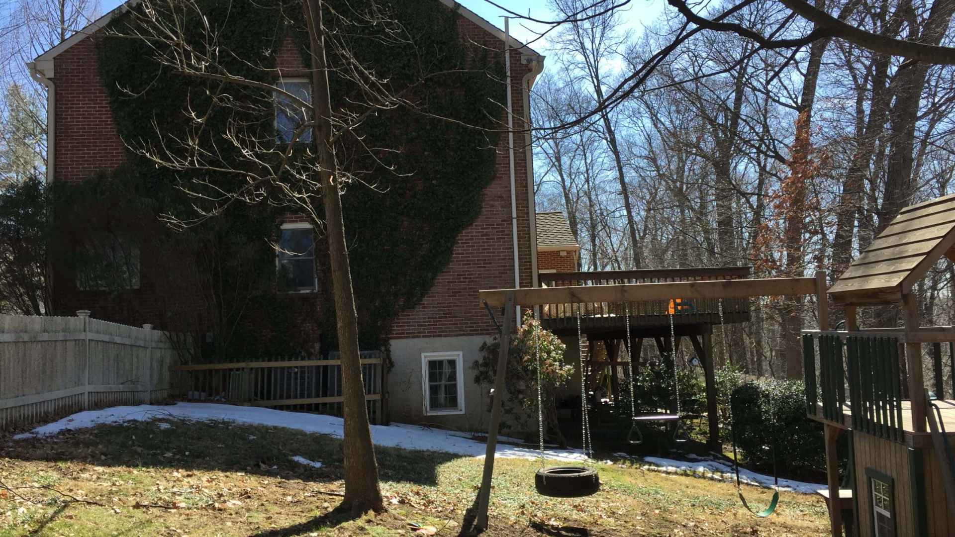 A backyard in Delaware County, PA, showcases a brick house adorned with ivy, a wooden deck with a roof, and a leafless tree. A swing hangs from the tree beside a wooden play structure. Patches of snow and scattered leaves enhance this charming outdoor living space under a clear blue sky.