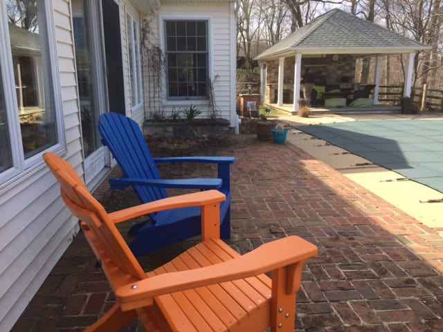 Two colorful Adirondack chairs, one orange and one blue, sit on a brick patio that showcases expert hardscaping services in Delaware County, PA. Nearby, a covered pool and a gazebo with seating beckon from the background. Leafless trees frame the scene, suggesting late fall or early spring.