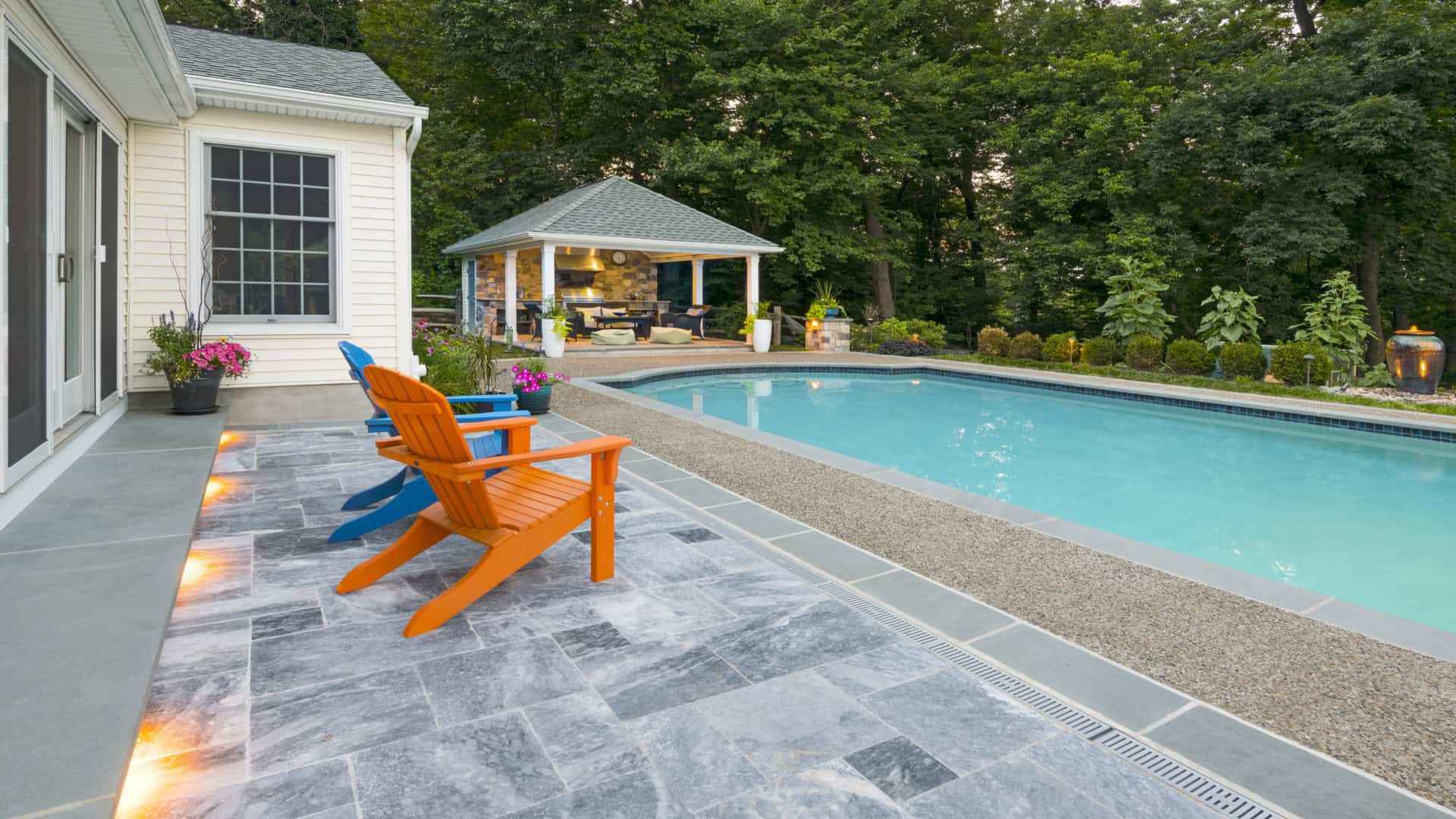 A serene backyard with a swimming pool surrounded by trees showcases expert landscaping services in Delaware County, PA. The stone patio features orange and blue Adirondack chairs. A cozy gazebo with seating is in the background, and warm lights illuminate the walkway for perfect outdoor living.
