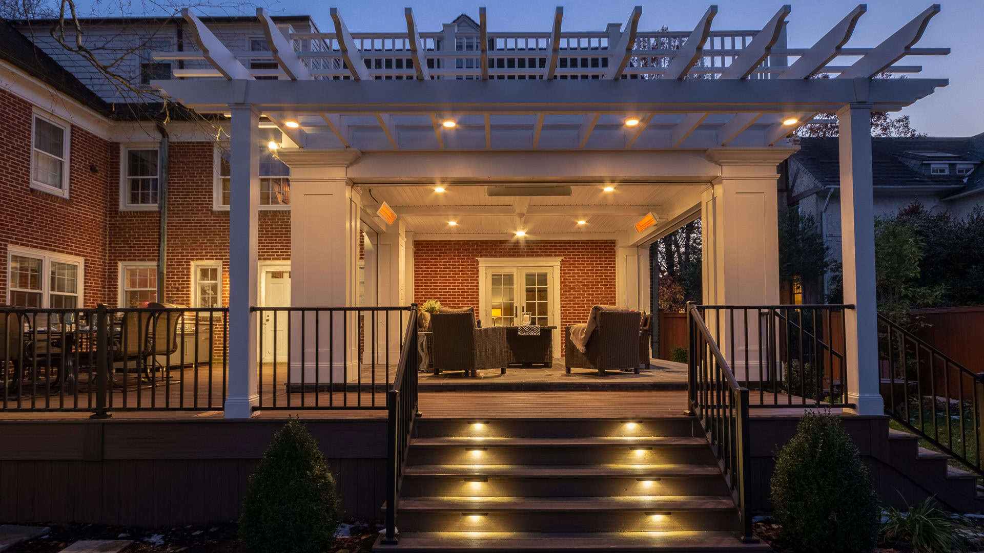 A well-lit patio attached to a brick house features a pergola with string lights, perfect for outdoor living in Delaware County, PA. Steps with embedded lights lead up to the area where cushioned chairs and a table are arranged under the pergola.