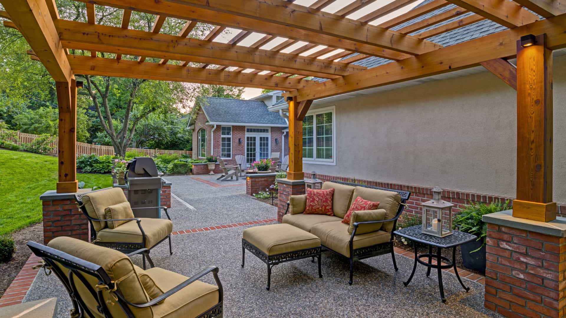 Enjoy a cozy outdoor living space in Delaware County, PA, featuring a covered patio with cushioned metal chairs and a small table under a charming wooden pergola. Nearby, the grill rests on a brick and gravel patio against the backdrop of a brick house surrounded by lush trees and lawn.