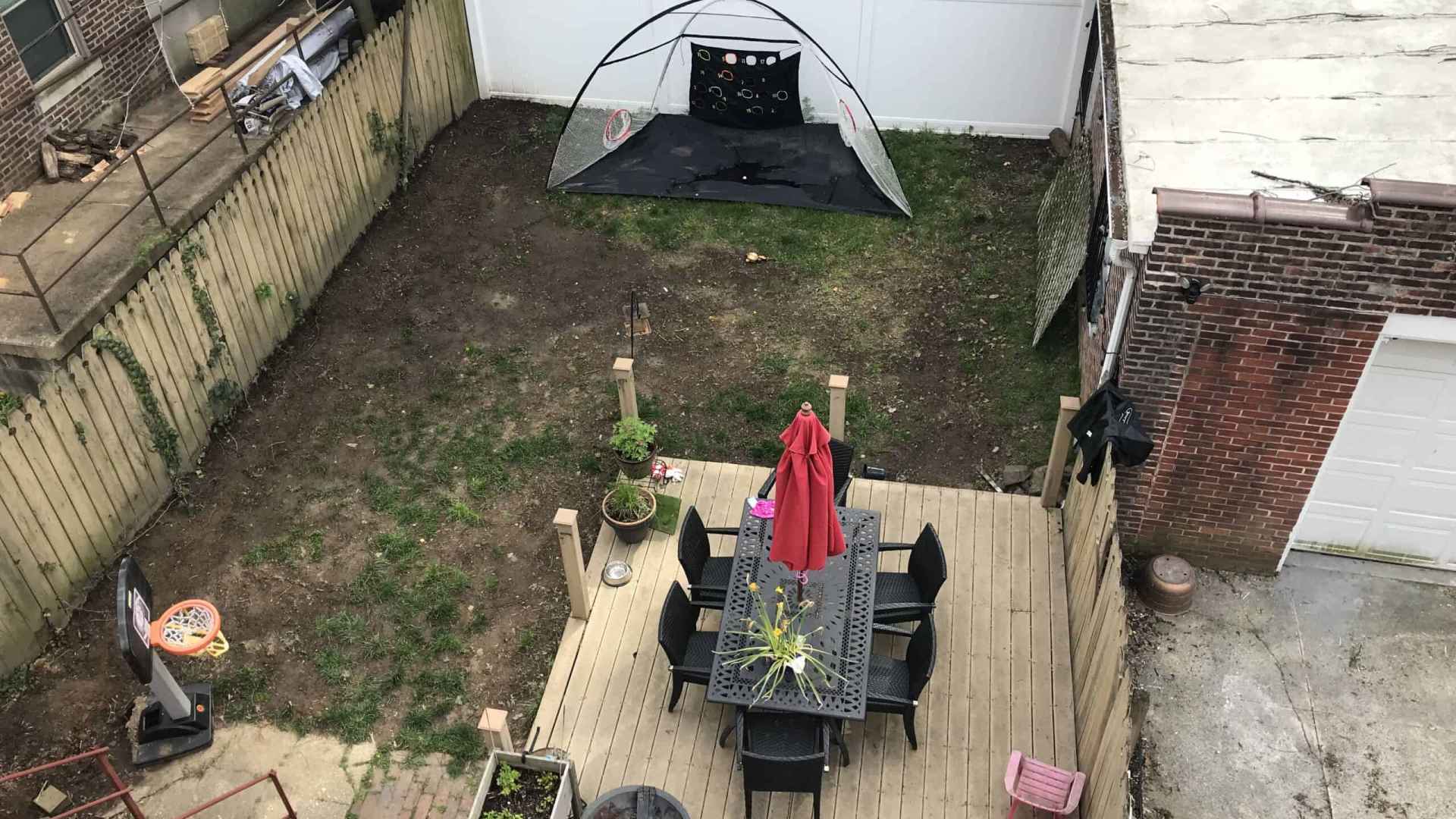 Aerial view of a backyard in Delaware County, PA, showcasing outdoor living with a wooden deck featuring a dining table, chairs, and a red umbrella. A soccer goal is set on the grass. Potted plants add charm, while a white fence and brick garage frame the space—a testament to fine landscaping services.