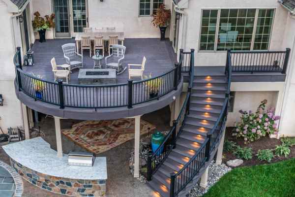 Aerial view of a two-story patio with a circular upper deck featuring a dining table, chairs, and fire pit. Curved staircase with glowing steps leads to the lower area with a bar and stone flooring, exemplifying premier outdoor living in Delaware County, PA. Vibrant green lawn in the foreground.