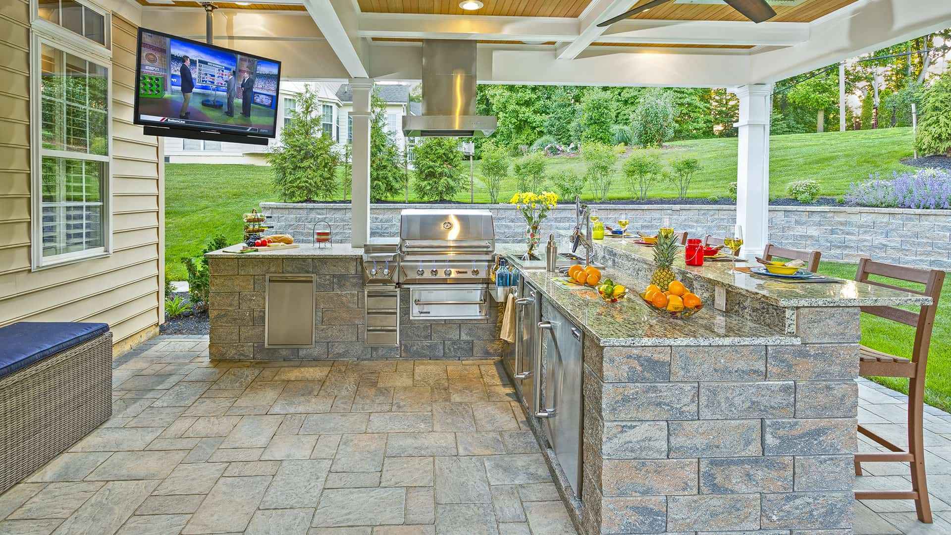 Outdoor living in Delaware County, PA offers a stunning kitchen equipped with a grill, oven, and granite countertop bar. A TV is mounted above the grill. Enjoy stone flooring surrounded by lush greenery and a retaining wall, perfect for hosting with fruits and drinks ready on the counter.