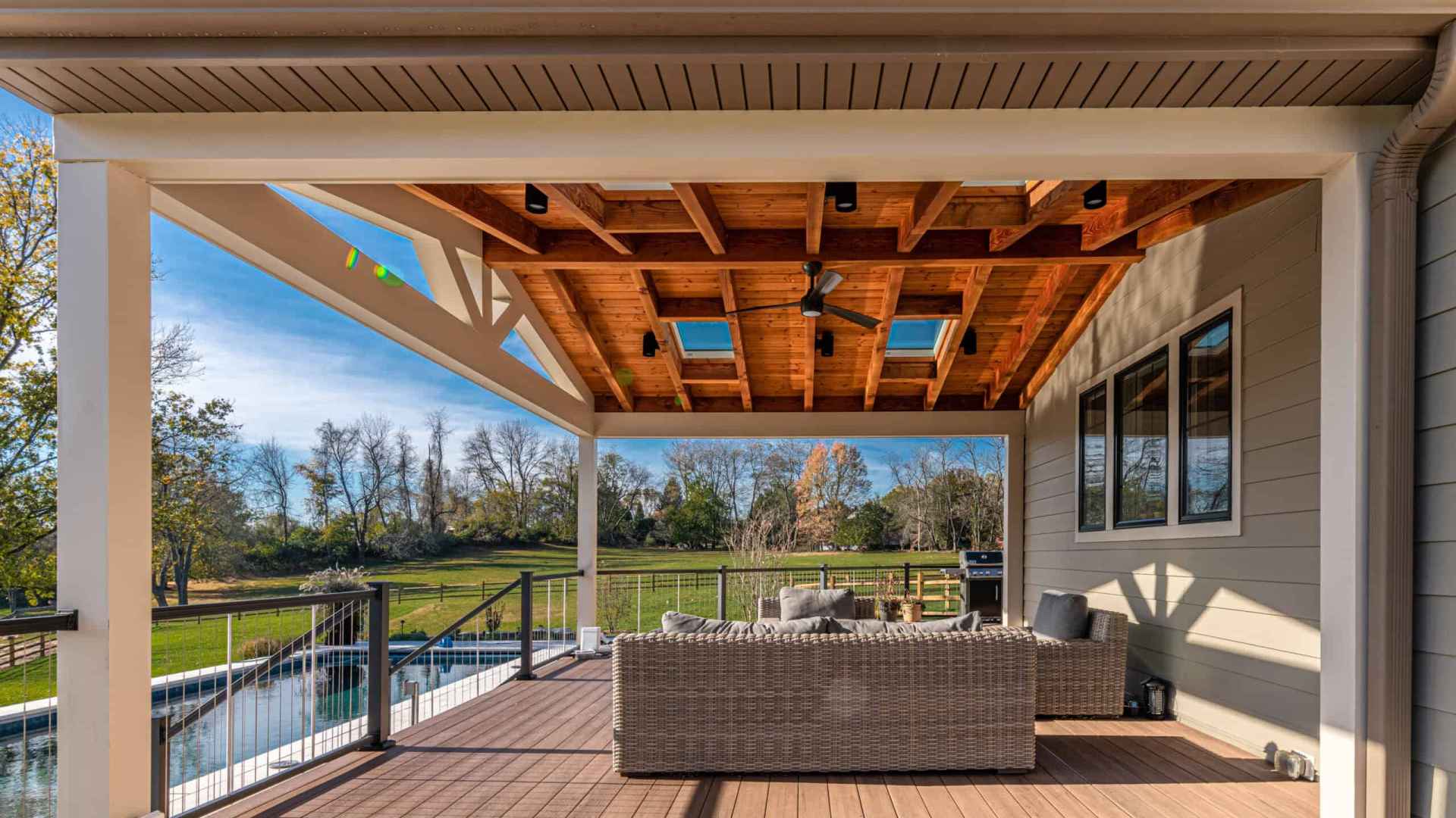Experience outdoor living at its finest on a covered patio with a wooden ceiling and skylights. Wicker seating graces the wooden deck as you overlook the grassy yard and fenced-in pool beneath a clear blue sky. Perfect for those seeking exceptional landscaping services in Delaware County, PA.