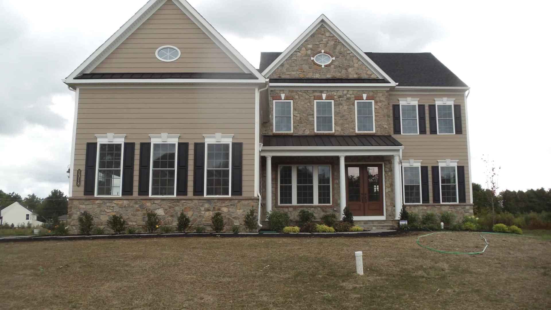 A two-story house with beige siding, stone accents, and white trim. It features dark shutters and multiple windows. The yard, perfect for outdoor living in Delaware County PA, has a patch of grass, shrubs near the house, and a garden hose on the lawn beneath a cloudy sky.