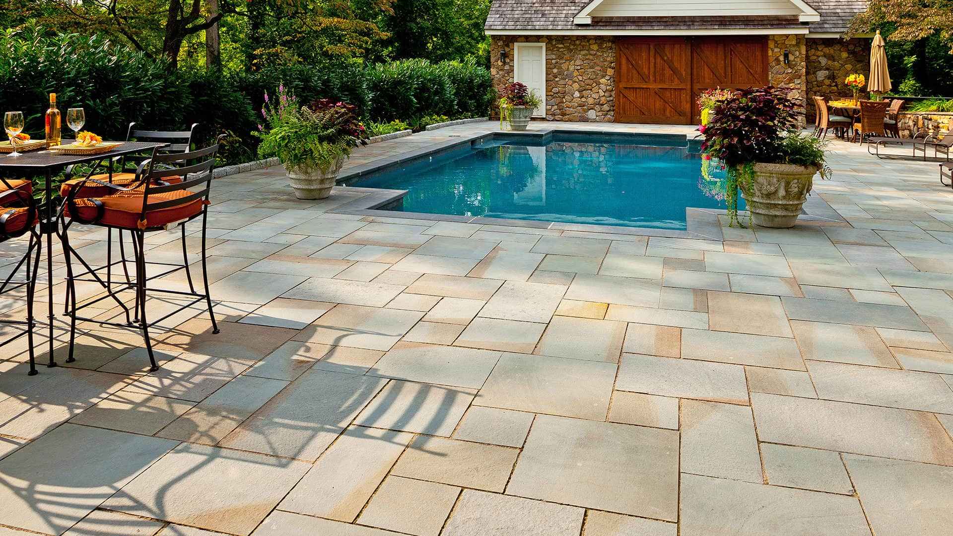 A backyard designed for outdoor living in Delaware County, PA, features a swimming pool surrounded by stone tiles. A table set with glassware and chairs with orange cushions adds charm, while lush greenery and a stone building with wooden doors complete the serene setting.
