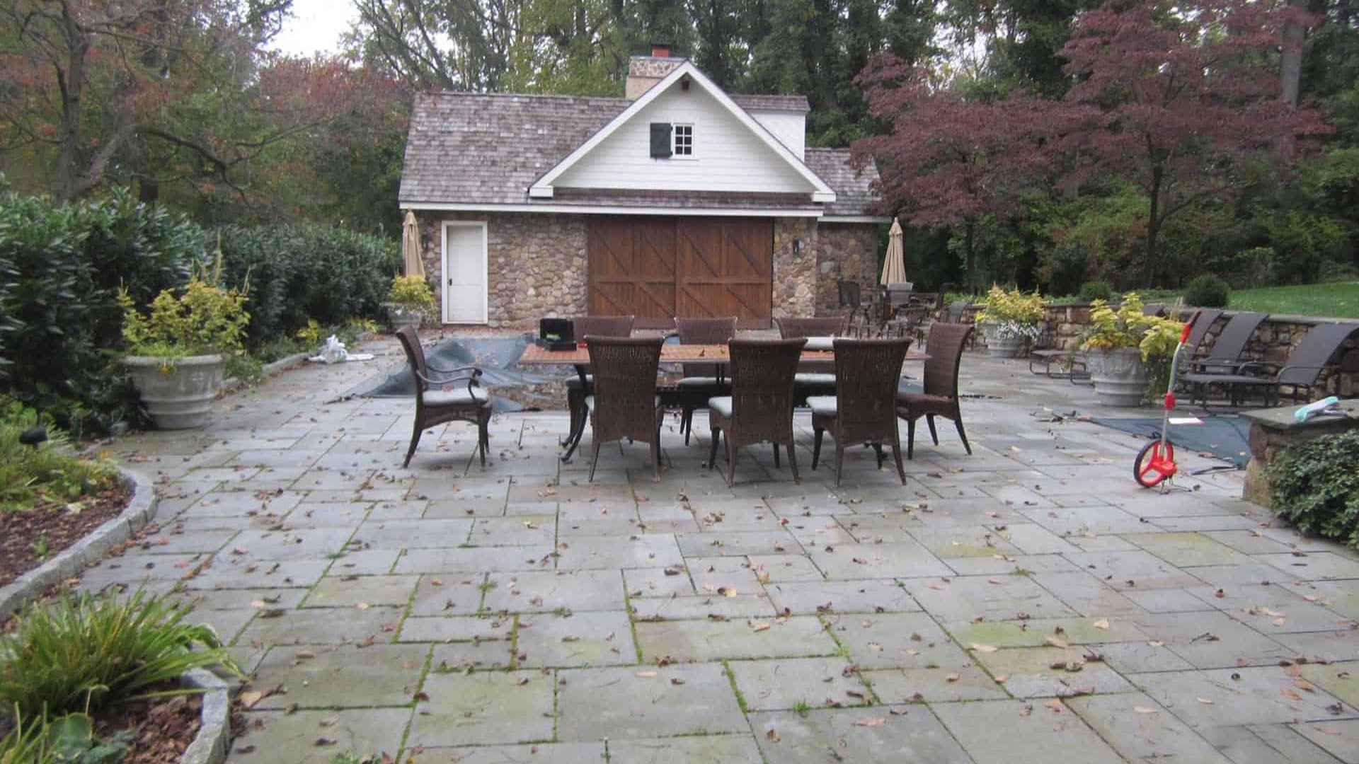 A stone patio features a spacious outdoor dining table with eight chairs, perfect for enjoying the beauty of autumn foliage. A wooden shed with a stone exterior adds charm, all framed by expert hardscaping services in Delaware County, PA. To the right, a child's toy lies on the lush lawn.