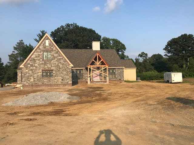 A stone house under construction on a clear day features a visible wooden framework at the entrance. The foreground is prepped for landscaping services in Delaware County, PA, with piles of gravel. Trees and a trailer are in the background, along with a shadow of the photographer.