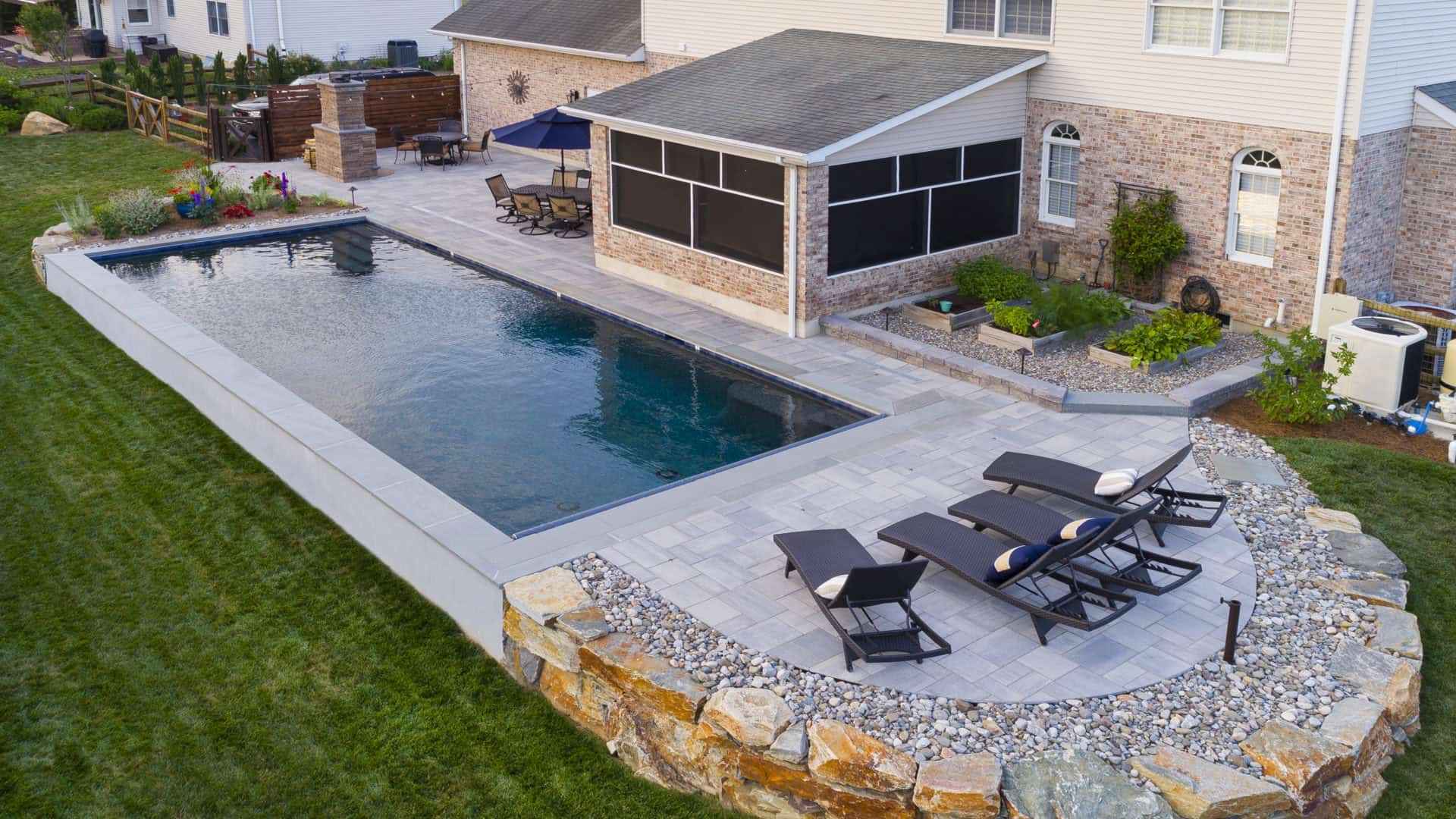 Aerial view of a backyard with a rectangular pool surrounded by a stone patio. Three black lounge chairs with cushions face the water. Nearby, there's a covered patio area connected to the brick house. Expert landscaping services in Delaware County, PA provide lush grass and stone borders.
