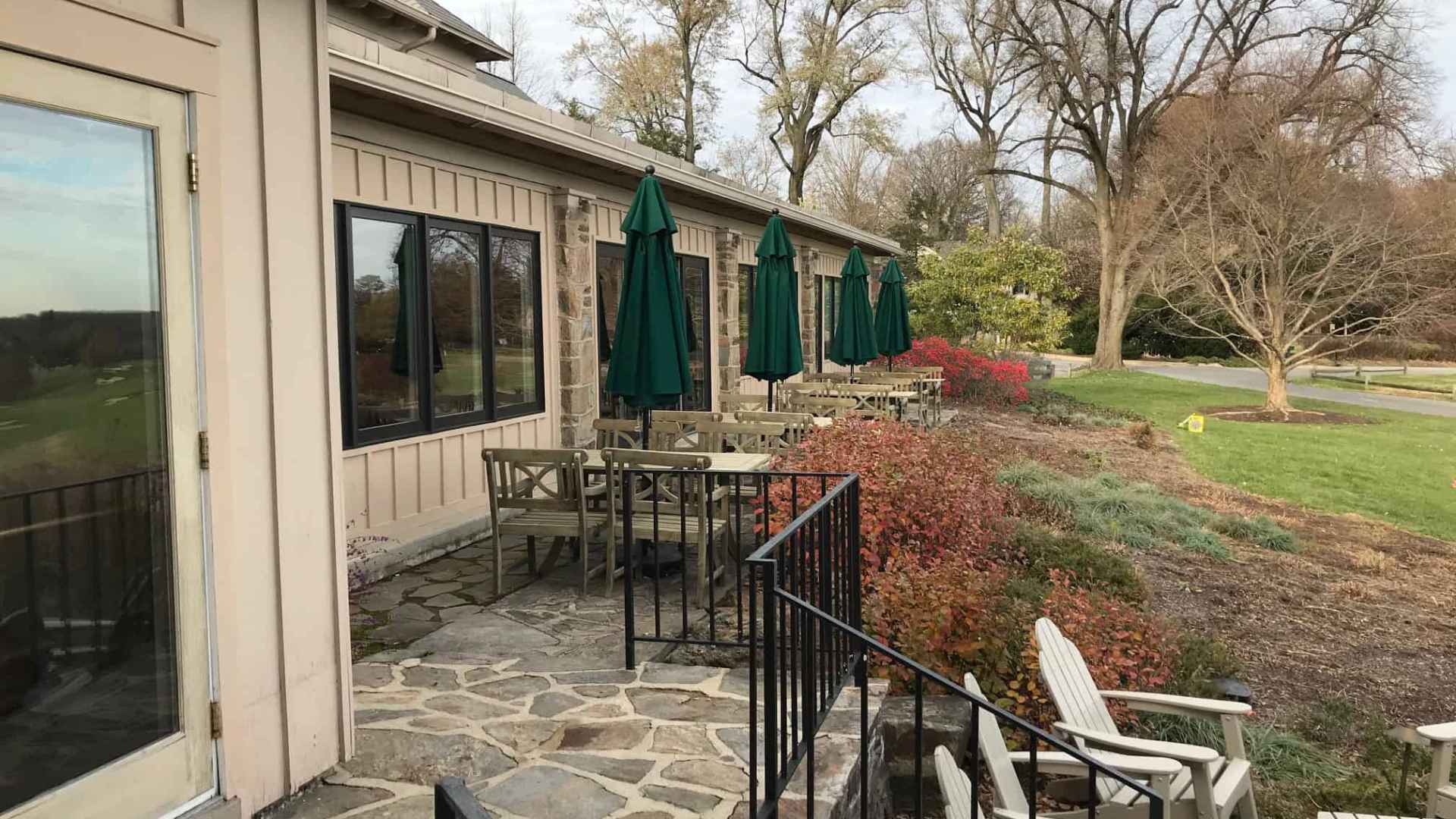 Outdoor patio with empty tables and closed green umbrellas at a stone building, exemplifying outdoor living Delaware County PA. A stone pathway leads to the patio with chairs, surrounded by dormant shrubs and trees. The sky is overcast.