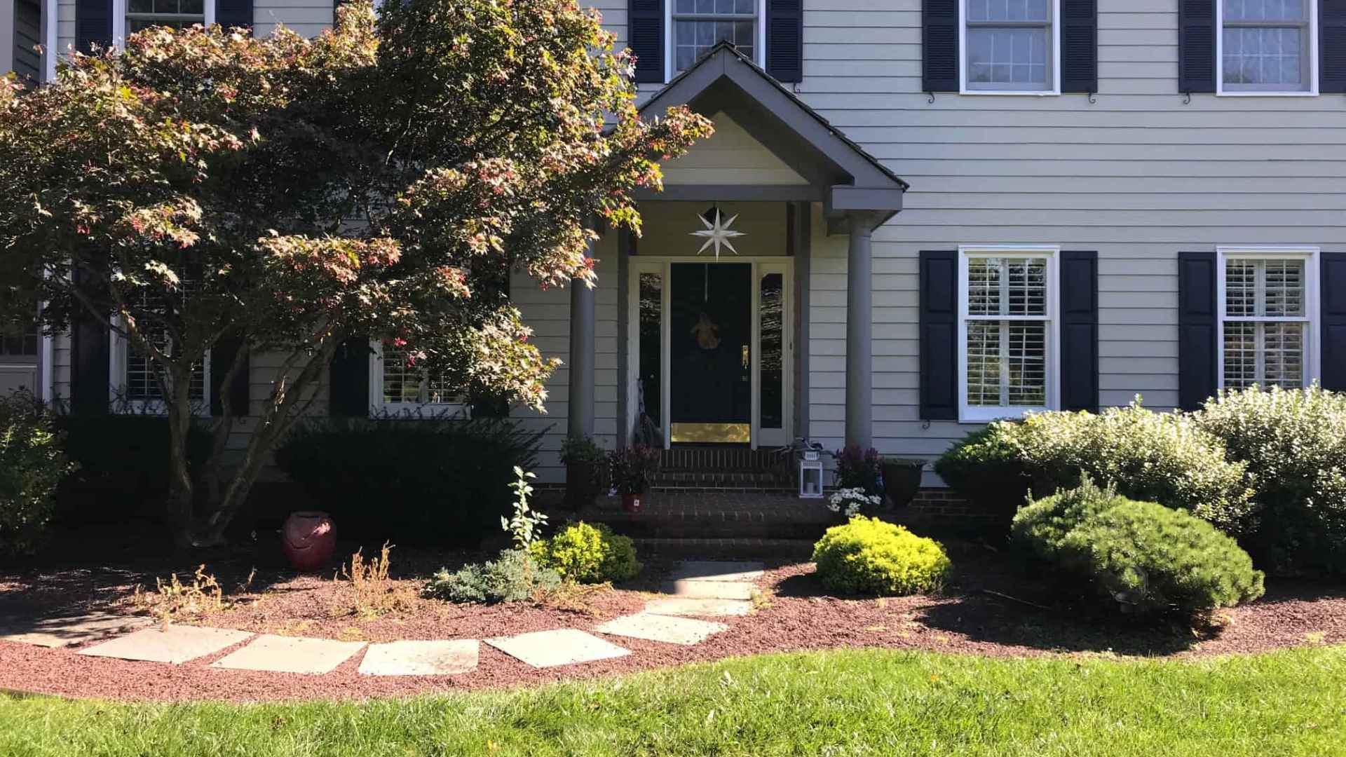 A two-story house with white siding and navy blue shutters, featuring a front porch with a small overhang. A curved pathway leads to the entrance, surrounded by a manicured garden with shrubs and a large tree, all enhanced by expert landscaping services in Delaware County, PA.