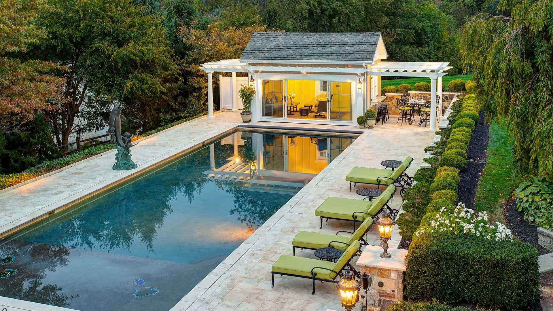 A luxurious backyard pool area with a reflective, rectangular pool surrounded by greenery showcases the elegance of premium landscaping services in Delaware County PA. Lounge chairs with green cushions, a warmly lit pavilion, and a bronze statue enhance this serene and inviting outdoor living space.