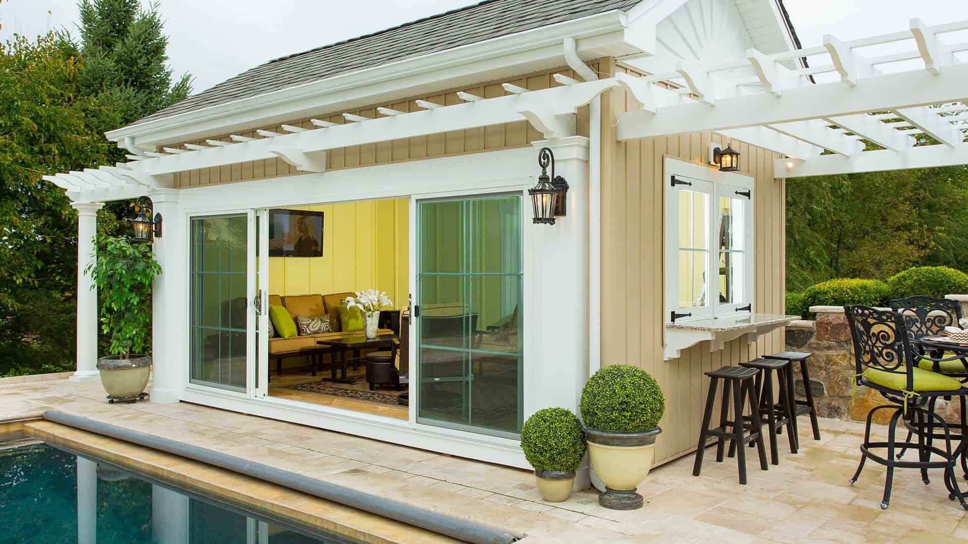 A small pool house with beige siding and a white pergola features sliding glass doors, a counter window, and outdoor seating. Decorative lanterns and potted plants add charm, enhancing the outdoor living experience in Delaware County PA. It's adjacent to a pool with a tiled deck, nestled in a green landscape.