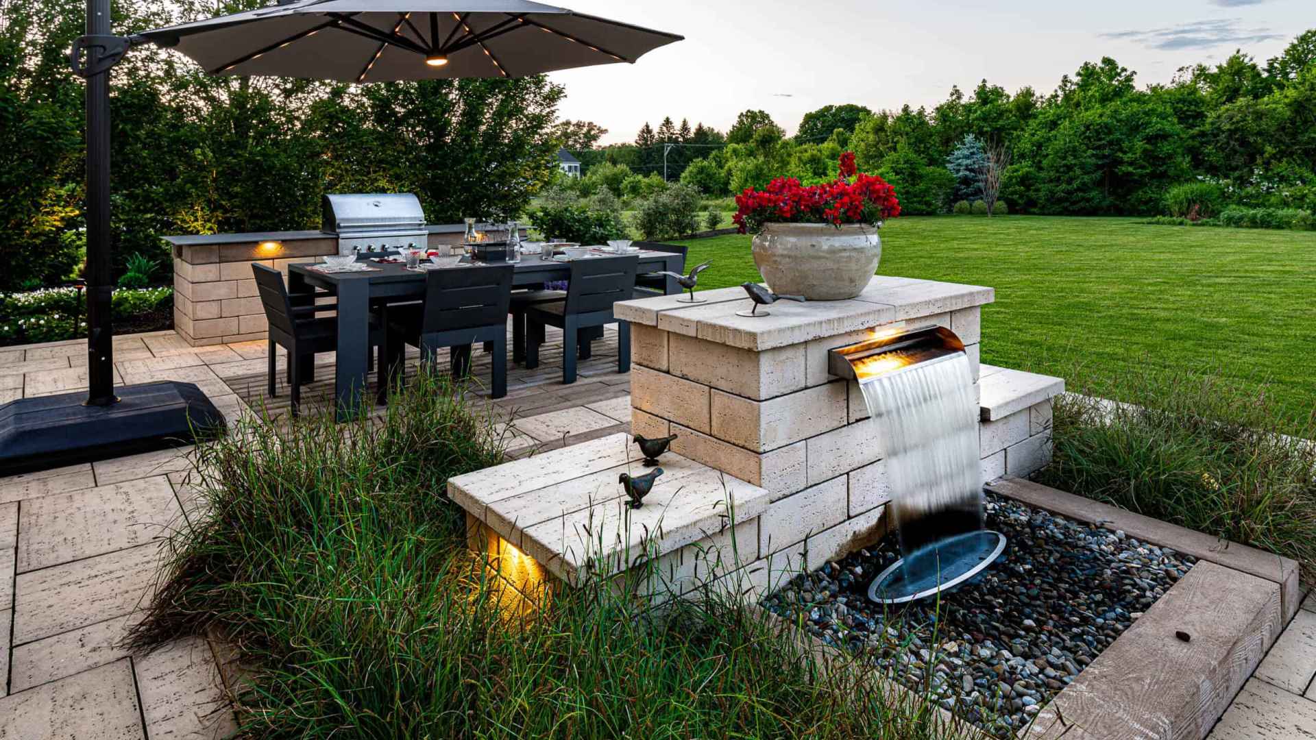 Explore the ultimate outdoor living in Delaware County, PA, with a patio featuring a dining table under a large umbrella. Nearby, a built-in grill and waterfall add charm. Surrounded by greenery, this space exemplifies expert hardscaping services in Delaware County, PA.