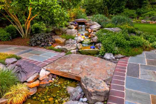 A landscaped garden features a small waterfall cascading over rocks into a pond. Surrounding greenery includes various plants and trees. The foreground showcases a colorful stone path bordered by bricks, a testament to expert landscaping services in Delaware County, PA, enhancing the serene natural scene.