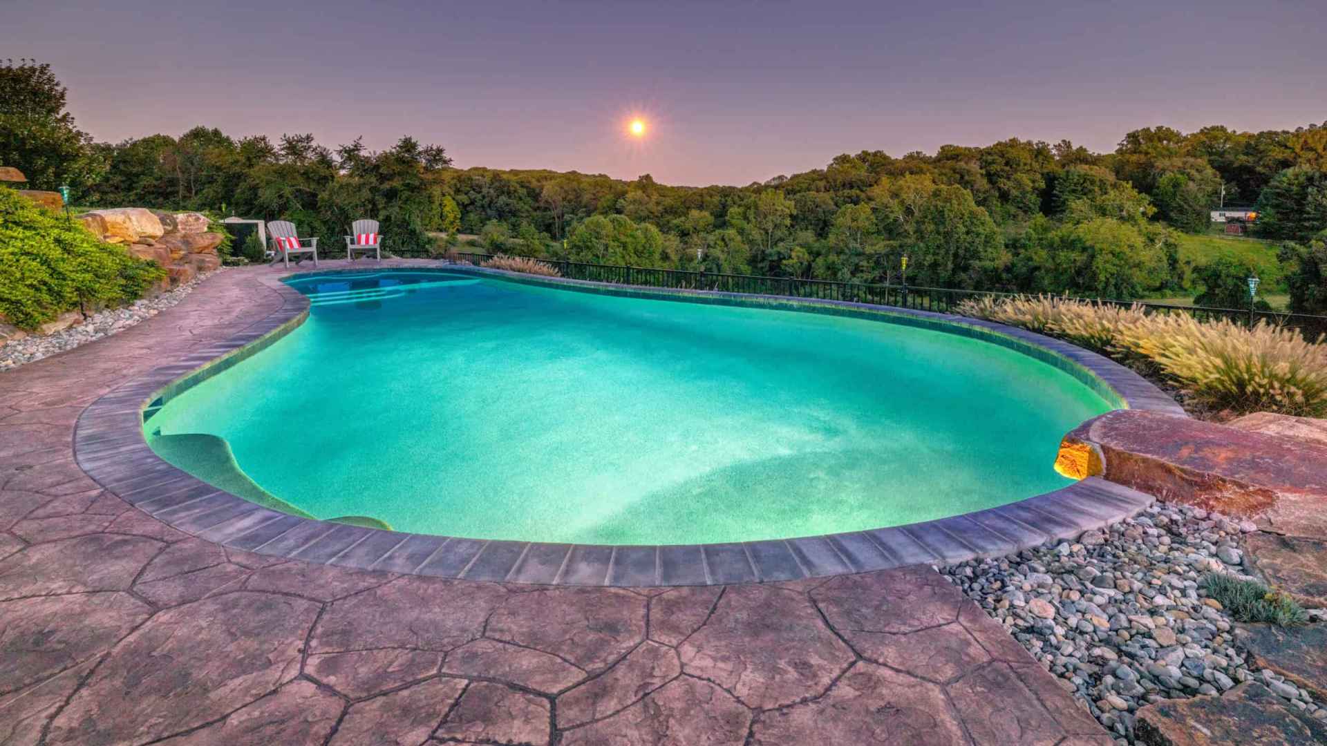 A tranquil scene of an outdoor pool with clear turquoise water, set amid expert hardscaping services in Delaware County, PA. Two red lounge chairs rest on the stone deck. Dense green trees and a bright moon in a dusky sky complete the picturesque outdoor living setting.