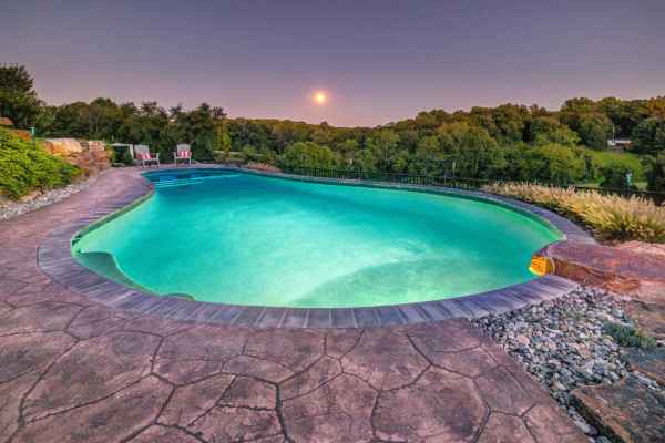 A tranquil scene of an outdoor pool with clear turquoise water, set amid expert hardscaping services in Delaware County, PA. Two red lounge chairs rest on the stone deck. Dense green trees and a bright moon in a dusky sky complete the picturesque outdoor living setting.