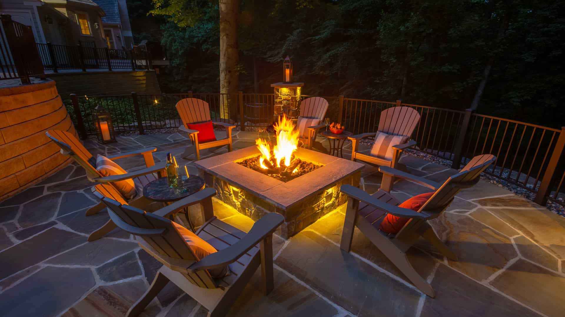 A cozy outdoor patio features a stone fire pit surrounded by six cushioned Adirondack chairs. Subtle lighting and the fire's glow illuminate the scene. With trees and a house in view, this serene evening ambiance highlights premium outdoor living in Delaware County, PA.