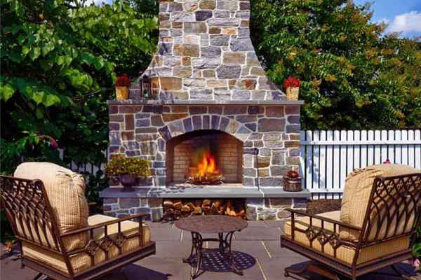 Outdoor living in Delaware County, PA, shines on this patio with a large stone fireplace at its center. A crackling fire warms two cushioned metal chairs and a small round table. Surrounded by lush trees and a white picket fence, it's the perfect blend of nature and design under a blue sky.
