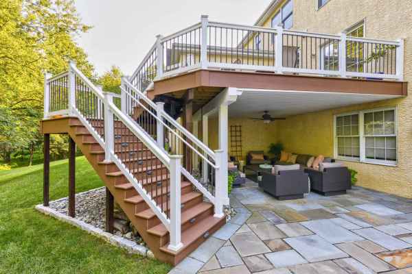 Outdoor view of a two-story deck featuring a staircase with white railings, leading to a ground-level patio. With wicker furniture and lush landscaping services Delaware County PA, this outdoor oasis is perfect for enjoying nature's beauty amidst the greenery and manicured lawn.