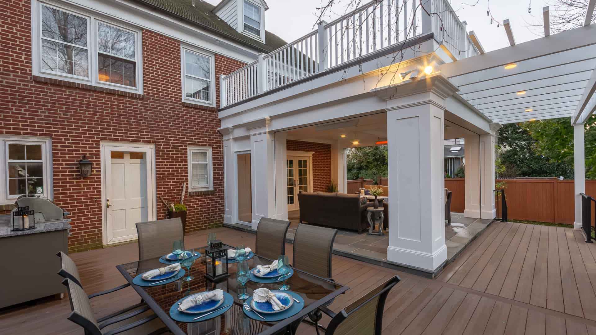 A spacious outdoor patio features a dining table set with blue and white dishware, chairs, a built-in grill, and a covered lounge area. Expertly crafted for outdoor living in Delaware County PA, the patio is attached to a brick house with large windows and a white balcony above.