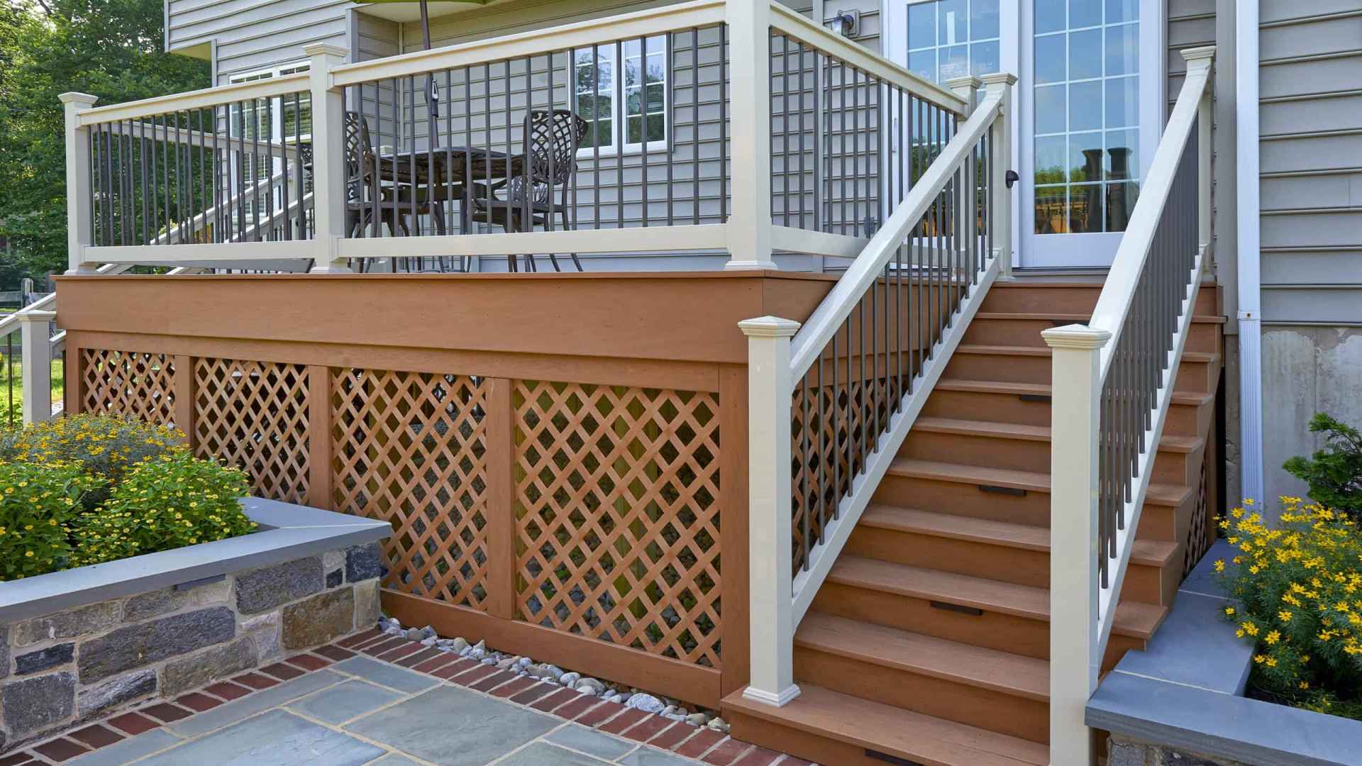 A wooden deck is attached to a house, featuring white railings and stairs leading down to a stone patio. Perfect for outdoor living in Delaware County, PA, it boasts a dining table with chairs and a green umbrella. Flowering plants add charm beside the stairs.