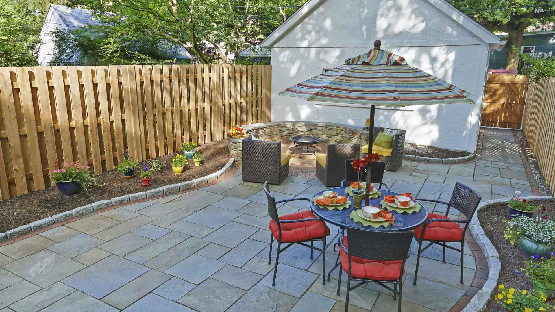 A backyard patio with a stone surface features a dining table with red cushioned chairs under a colorful striped umbrella. Nearby is a circular wicker seating area with a fire pit. This outdoor living space in Delaware County, PA, is beautifully surrounded by wooden fencing and lush greenery.