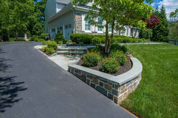 A well-maintained suburban home with gray stone exterior and white trim, nestled in Delaware County PA. The house is surrounded by lush greenery, a stone wall, manicured bushes, and colorful flower beds—all enhanced by expert landscaping services. A curved black asphalt driveway leads up to the garage.