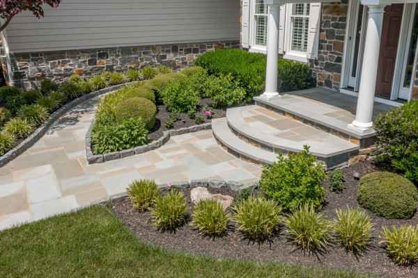 A well-maintained garden with a curved stone pathway leading to a house entrance showcases professional landscaping services in Delaware County, PA. Neatly trimmed bushes and small plants line the path and steps, while stone accents and white pillars frame the front door beautifully.